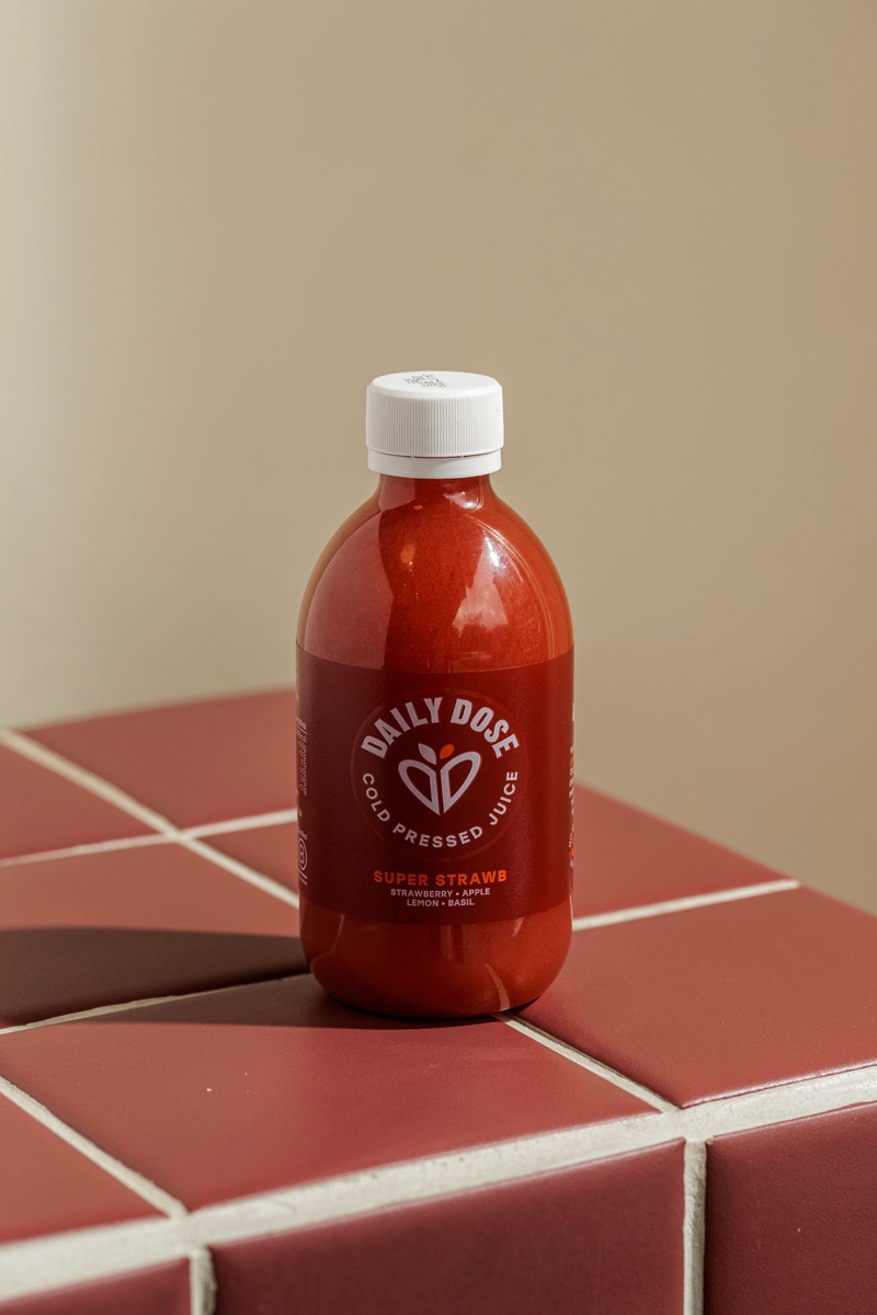 Daily Dose Super Strawb cold-pressed juice bottle on burgundy tile.