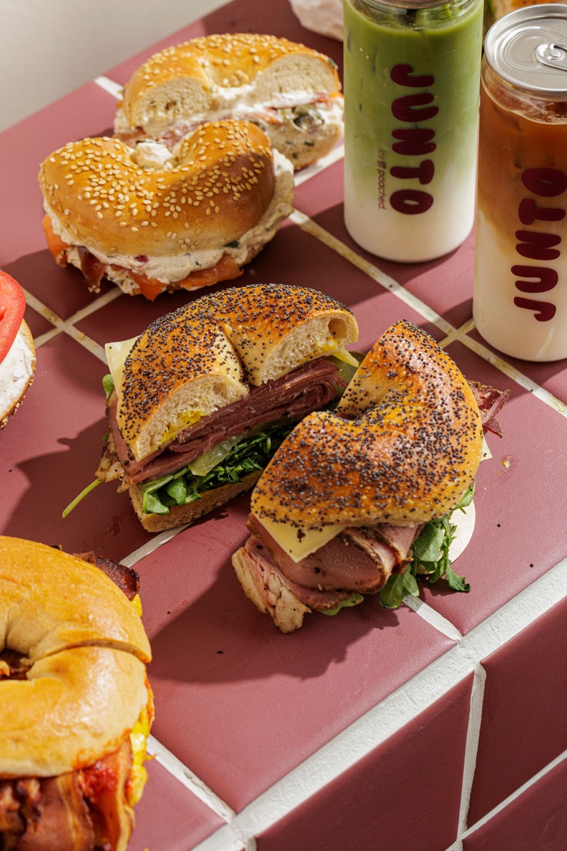 A spread of Junto bagels with JUNTO iced matcha and iced coffee cans on burgundy tile.