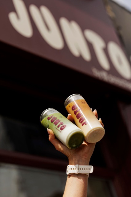 A hand lifting two JUNTO cans up under the burgundy JUNTO shopfront sign.