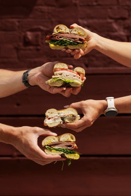 Four hands holding four halved bagels — pastrami, turkey, salmon cream cheese — against the burgundy wall.