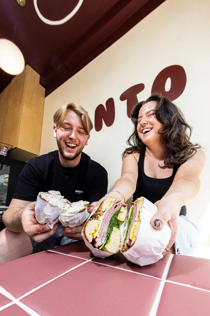 Two members of the team laughing as they hold up bagels.